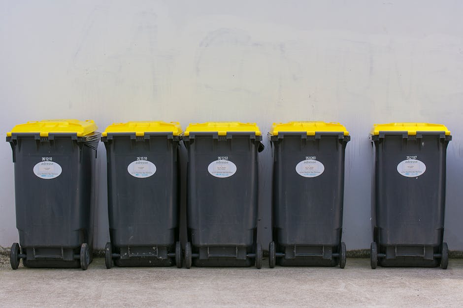 A collection of overflowing black, black-and-white, and red rubbish bins and open-top wheelie bins on a paved street, with numerous cardboard boxes, plastic bags, paper, and discarded packaging scatte