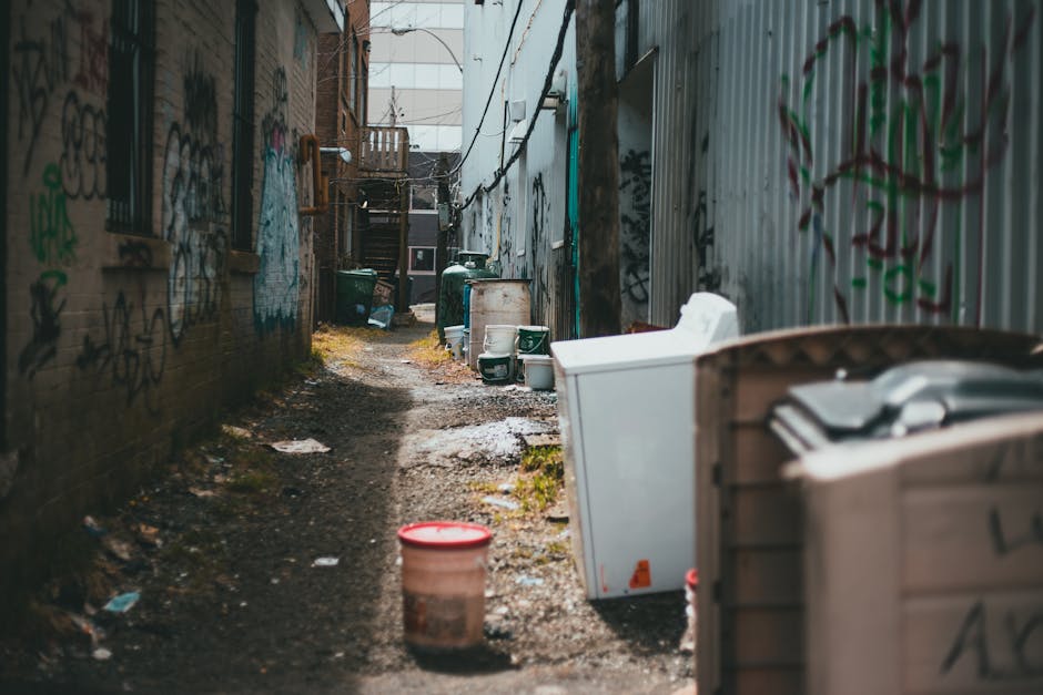 A large pile of black rubbish bags, some partially torn, stacked against a red metal door that is covered in graffiti and stickers. The rubbish includes cardboard boxes, flattened packaging, and misce